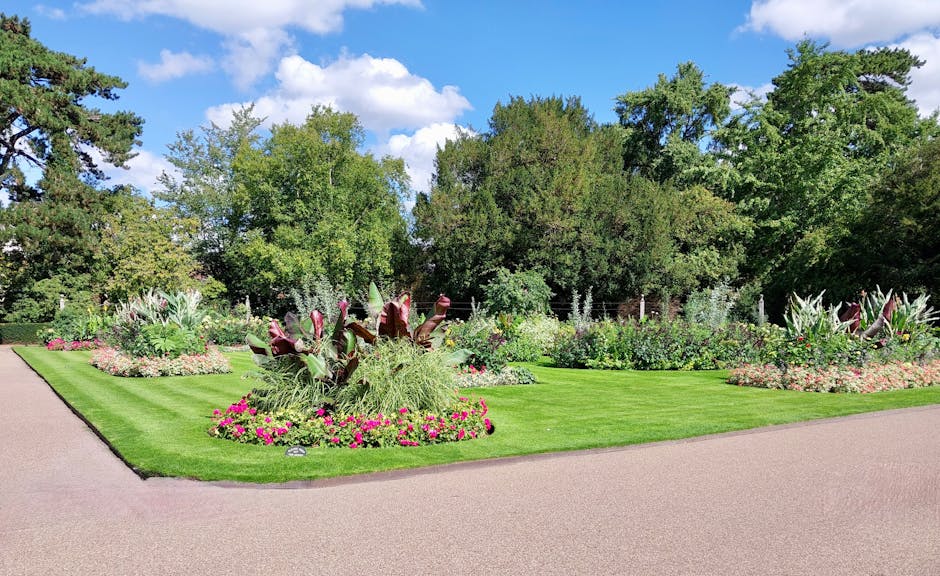 A landscaped garden featuring a well-maintained, vibrant green lawn bordered by flower beds filled with colorful plants and flowering shrubs. In the foreground, there are various ornamental plants wit